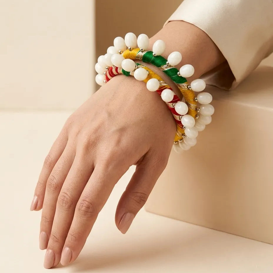 Woman's hand wearing stacked red yellow green silk resham thread bangles with white oval shell drops resting on cream surface
