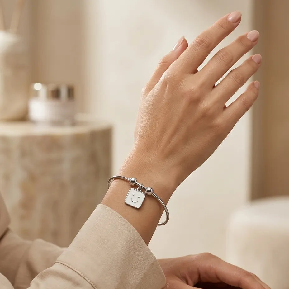 Woman wearing silver smiley face square enamel charm bangle bracelet on wrist with cream satin sleeve