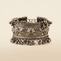Oxidized silver wide cuff bangle with embossed floral medallion panel rope-wire borders and cascading silver ghungroo ball drops on cream background