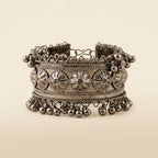 Oxidized silver wide cuff bangle with embossed floral medallion panel rope-wire borders and cascading silver ghungroo ball drops on cream background
