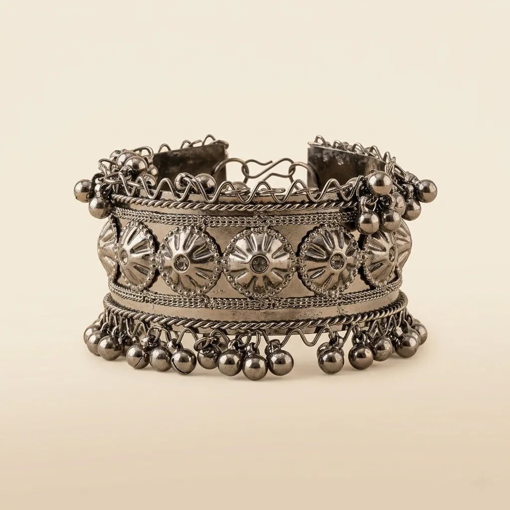 Oxidized silver wide cuff bangle with embossed floral medallion panel rope-wire borders and cascading silver ghungroo ball drops on cream background