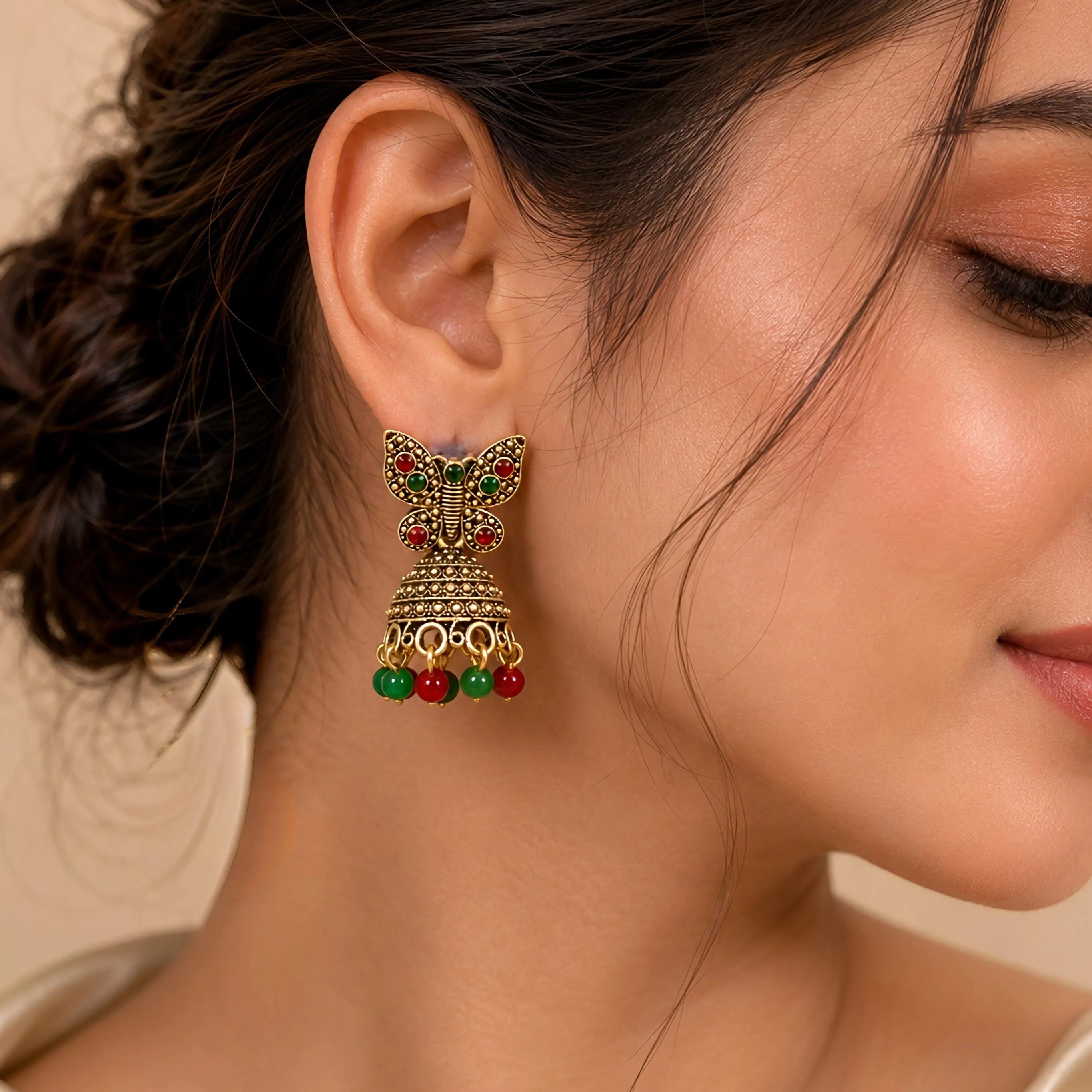 Close-up of a woman wearing gold butterfly earrings with red and green beads on a neutral background