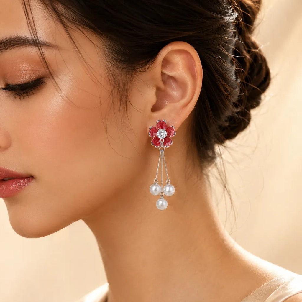 A close-up of a woman wearing a dangling flower earring with red gemstone petals, a clear center stone, and three dangling white pearls on a soft beige background.