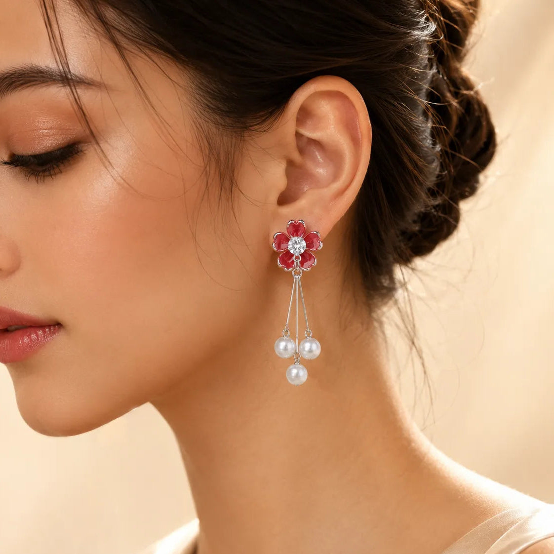 A close-up of a woman wearing a dangling flower earring with red gemstone petals, a clear center stone, and three dangling white pearls on a soft beige background.