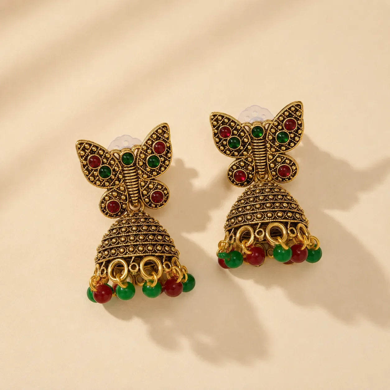 Antique gold butterfly jhumka earrings with red and green stone embellishments and bead drops on beige background