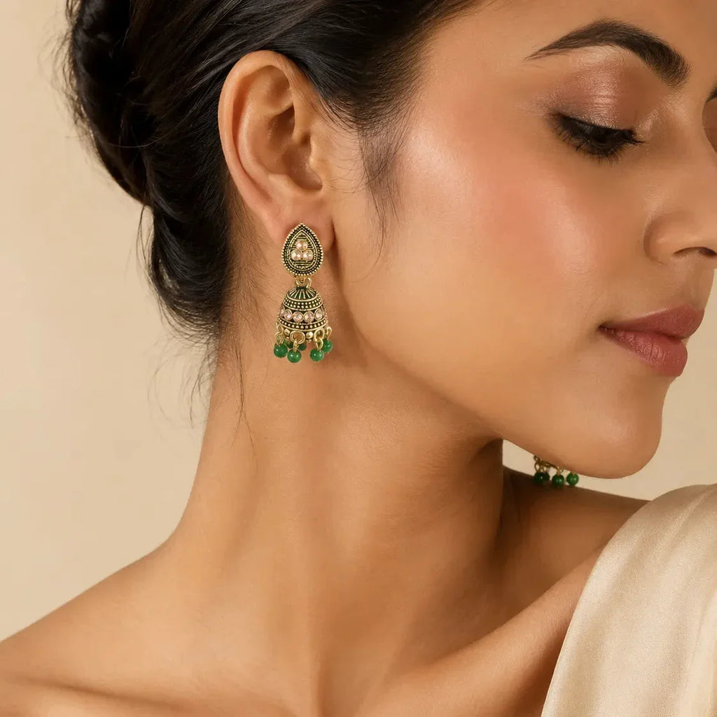 Woman wearing antique gold green meenakari teardrop jhumka earrings with emerald bead drops styled with cream satin outfit