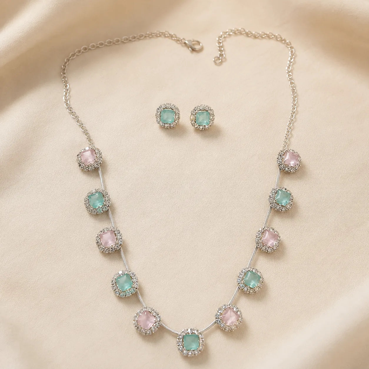 Silver collar necklace and stud earrings with alternating pink and mint cushion cut crystals, laid flat on beige background