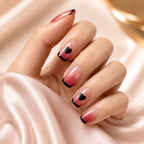 Woman's hand wearing SAVNI pink ombre press-on nails with black heart French tips, close-up manicure photo on satin fabric