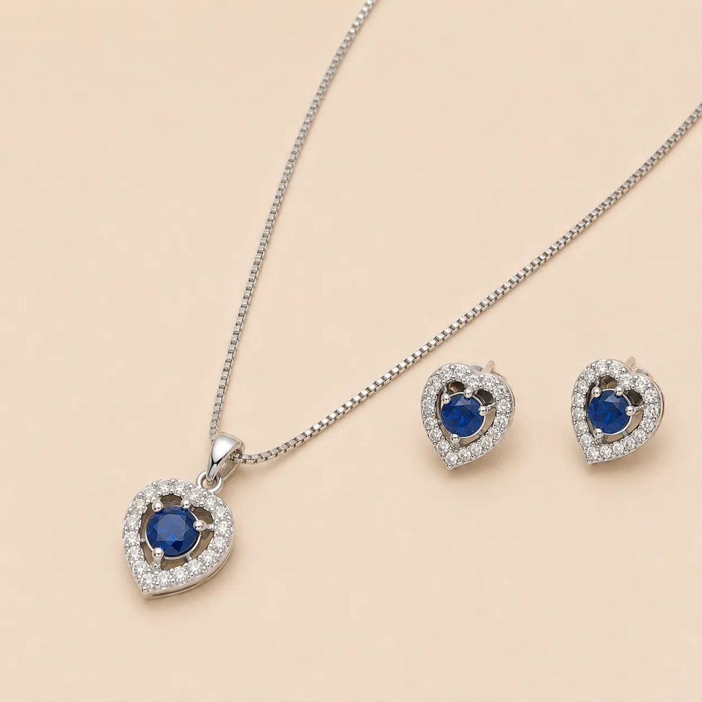 Close-up of silver heart shaped necklace pendant and earrings with blue gemstone centers on beige backdrop