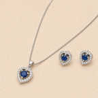 Close-up of silver heart shaped necklace pendant and earrings with blue gemstone centers on beige backdrop