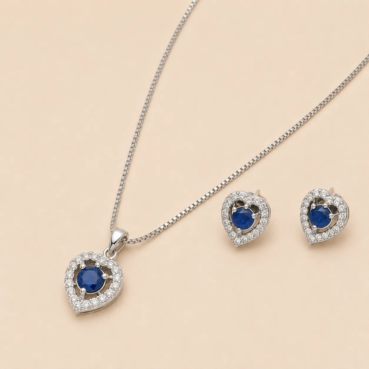 Close-up of silver heart shaped necklace pendant and earrings with blue gemstone centers on beige backdrop