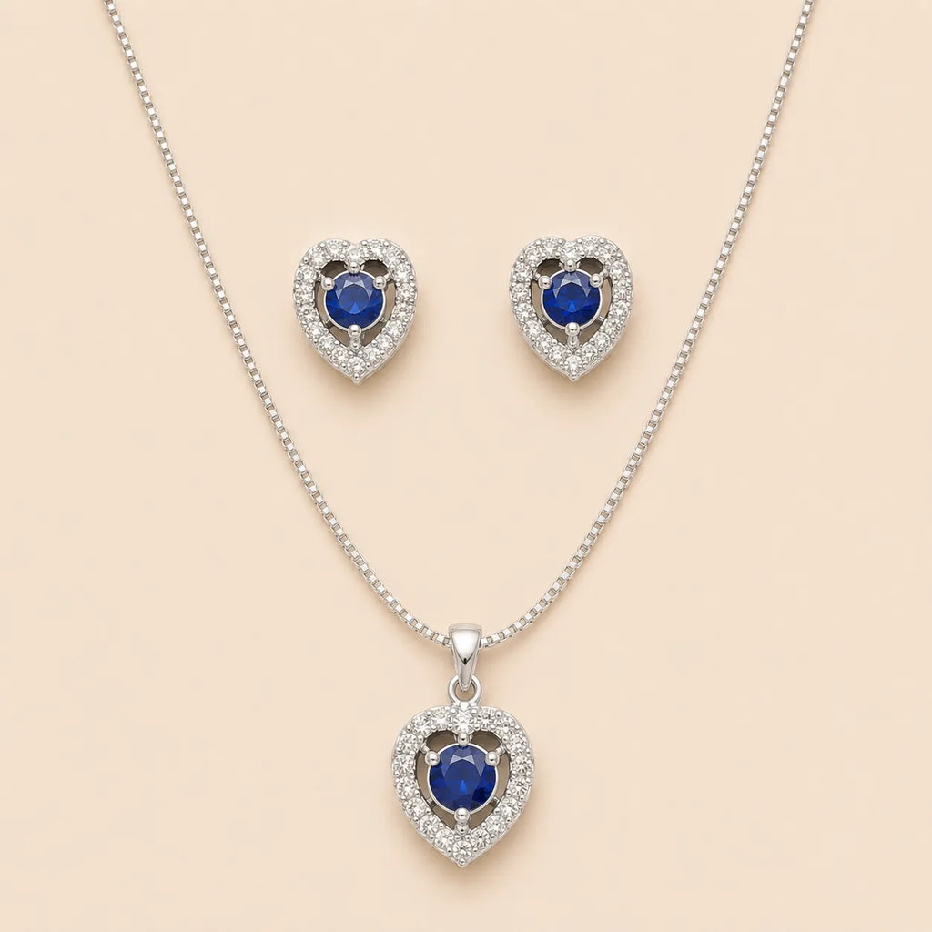 Silver heart halo necklace and stud earrings set with blue sapphire-style center stones, laid flat on beige background