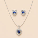 Silver heart halo necklace and stud earrings set with blue sapphire-style center stones, laid flat on beige background