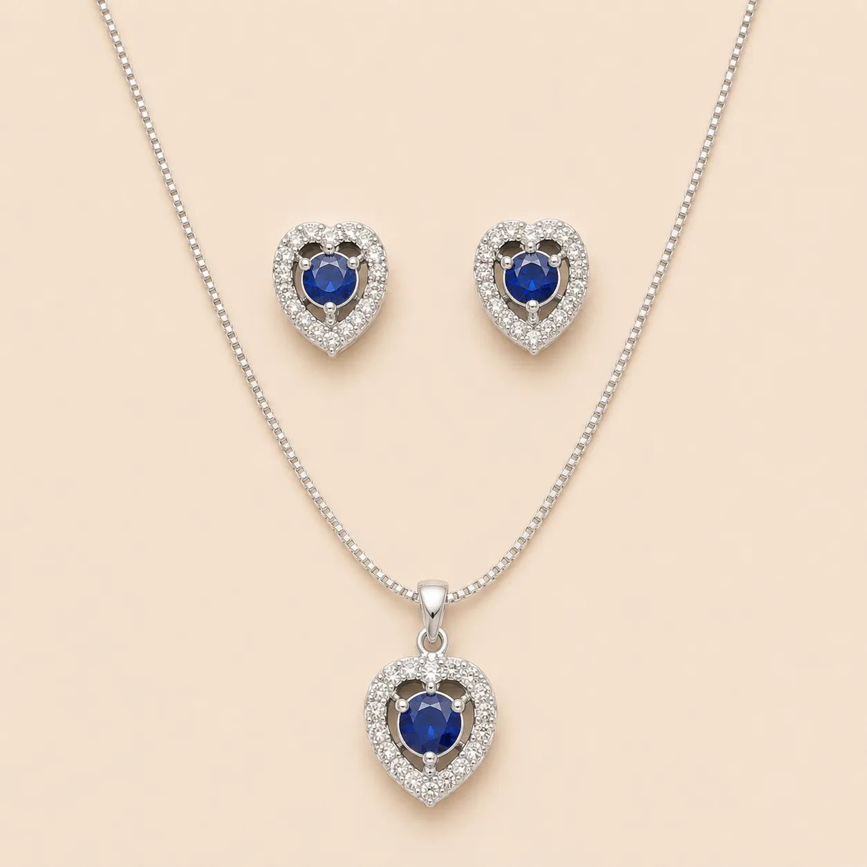 Silver heart halo necklace and stud earrings set with blue sapphire-style center stones, laid flat on beige background
