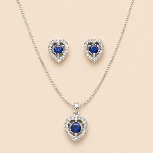 Silver heart halo necklace and stud earrings set with blue sapphire-style center stones, laid flat on beige background