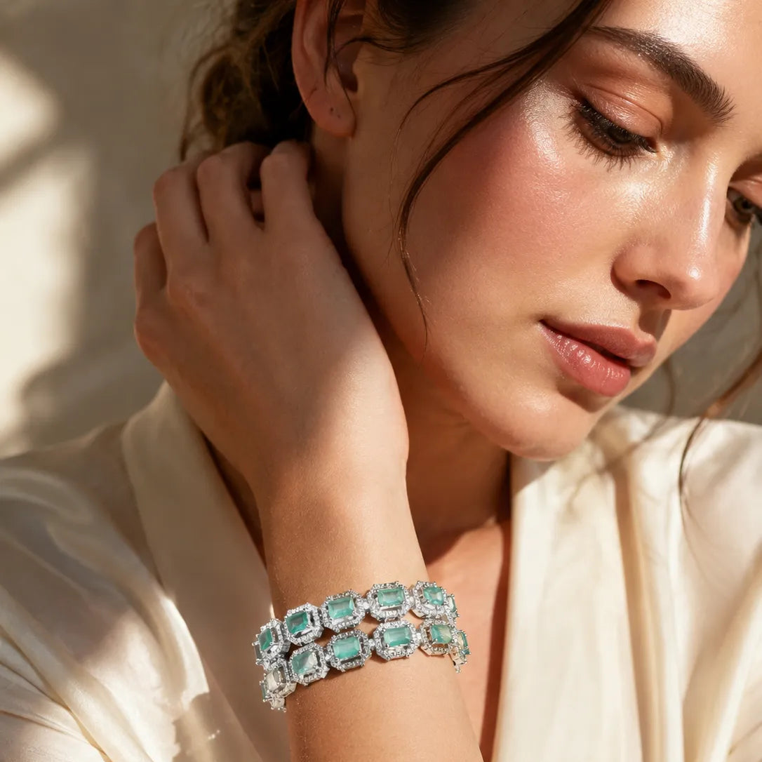 Woman wearing two silver mint green emerald cut halo CZ bangle bracelets stacked on wrist with cream satin outfit in sunlight