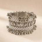 Silver white enamel wide cuff bangle with embossed checkerboard motifs and double-row silver ghungroo ball drop chains standing upright on beige background