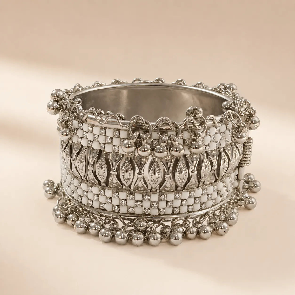 Silver white enamel wide cuff bangle with embossed checkerboard motifs and double-row silver ghungroo ball drop chains standing upright on beige background