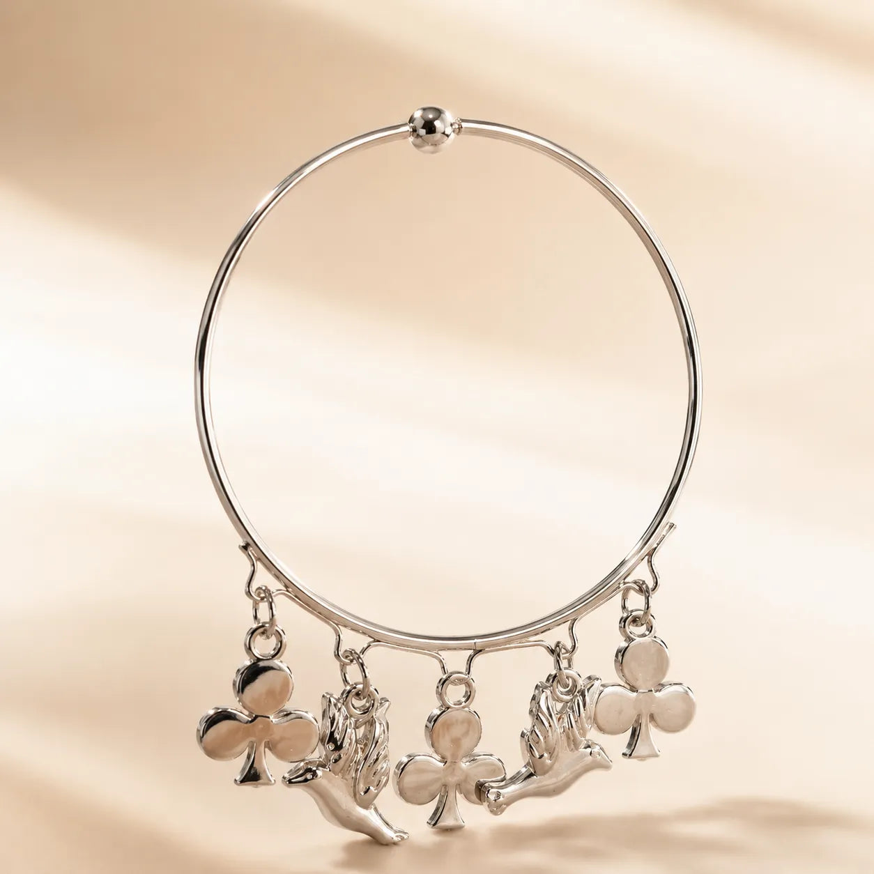 Silver slim wire bangle bracelet with five alternating angel cherub and club suit charms hanging from lower arc on beige background